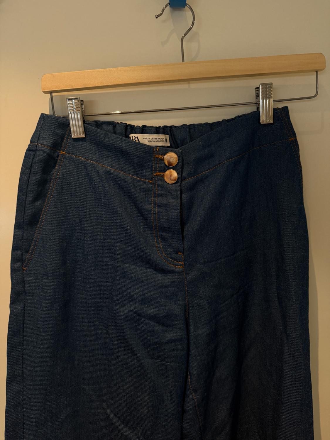 Dark blue denim trousers hanging on a wooden hanger, with two brown buttons and visible orange stitching.