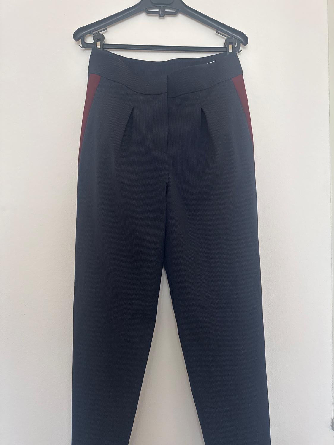 Black trousers with red details on the sides, hanging on a black hanger against a white wall.