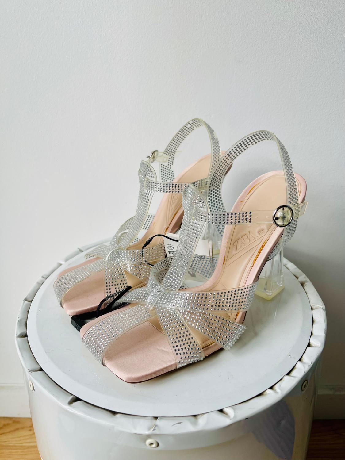 Clear heeled sandals with sparkly silver straps, on a white stool. Visible text: "ZARA".