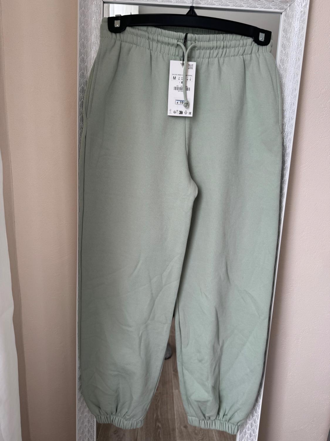 Light green tracksuit bottoms hanging on a black hanger in front of a mirror, with a size M label visible.
