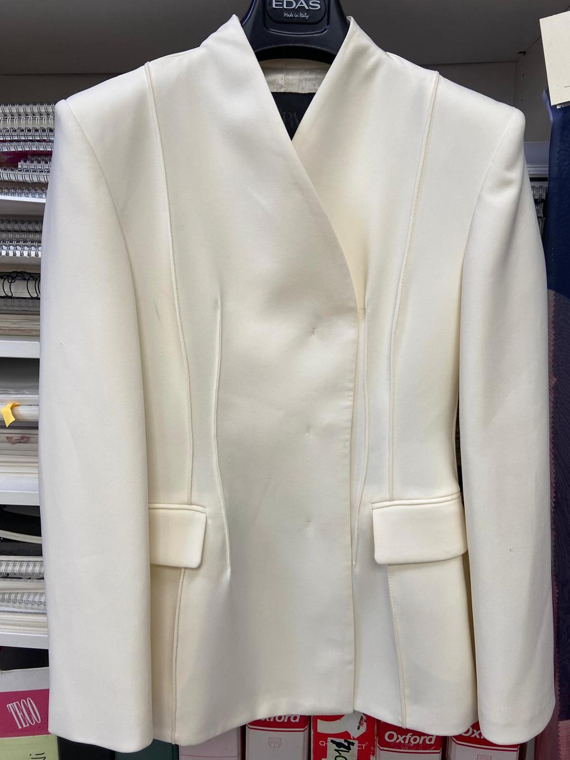 Structured white jacket hanging on a hanger with the text “EDAS Made in Italy”. Background with shelves and boxes bearing the text “TECO” and “Oxford”.