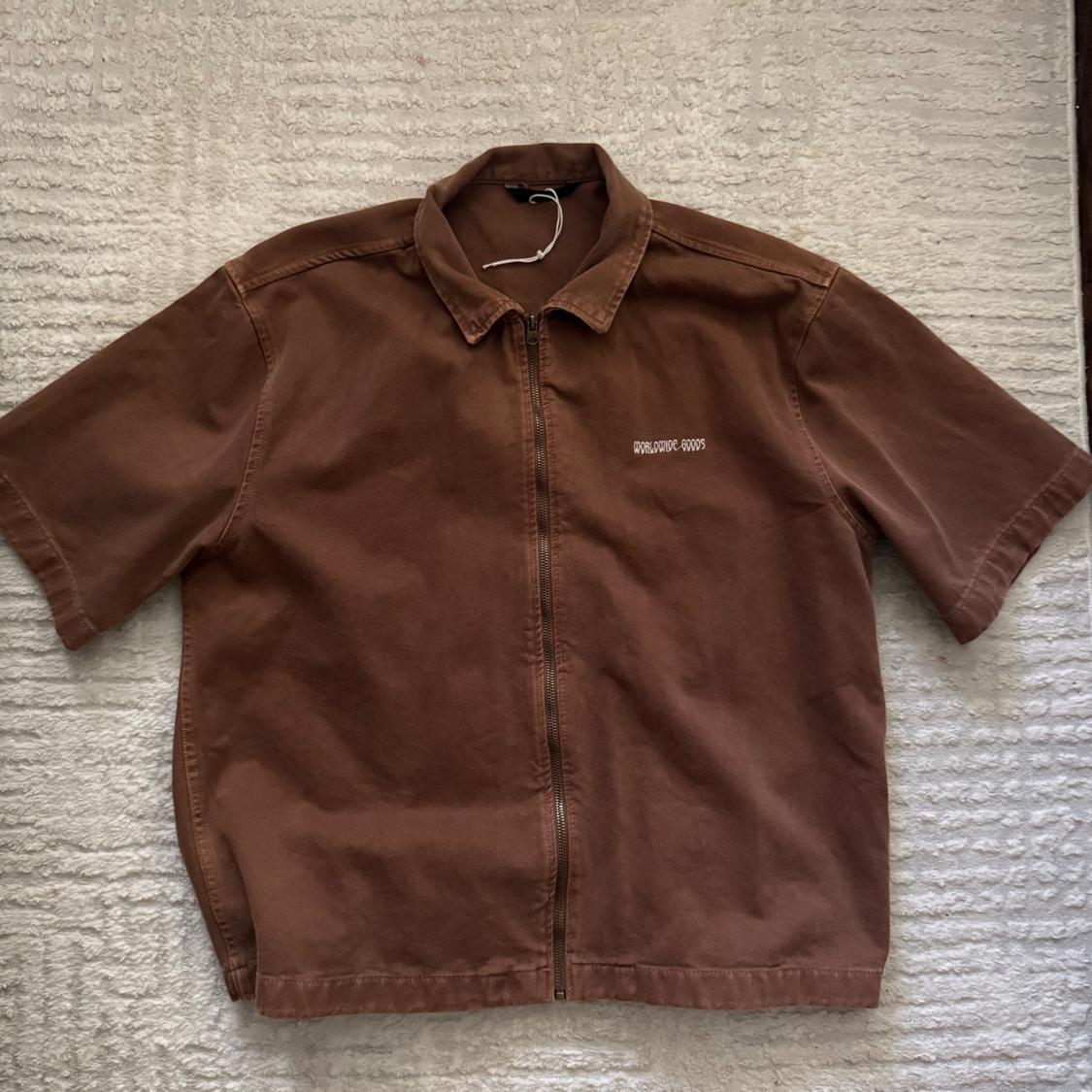 Brown short-sleeve shirt with a zipper and the text WORLDWIDE GOODS on the chest, laid out on a light-colored rug.