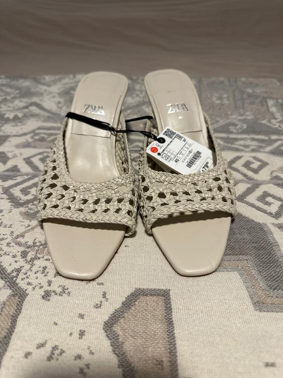 Beige low-heeled sandals with a braided design and price tag on a geometric patterned surface.