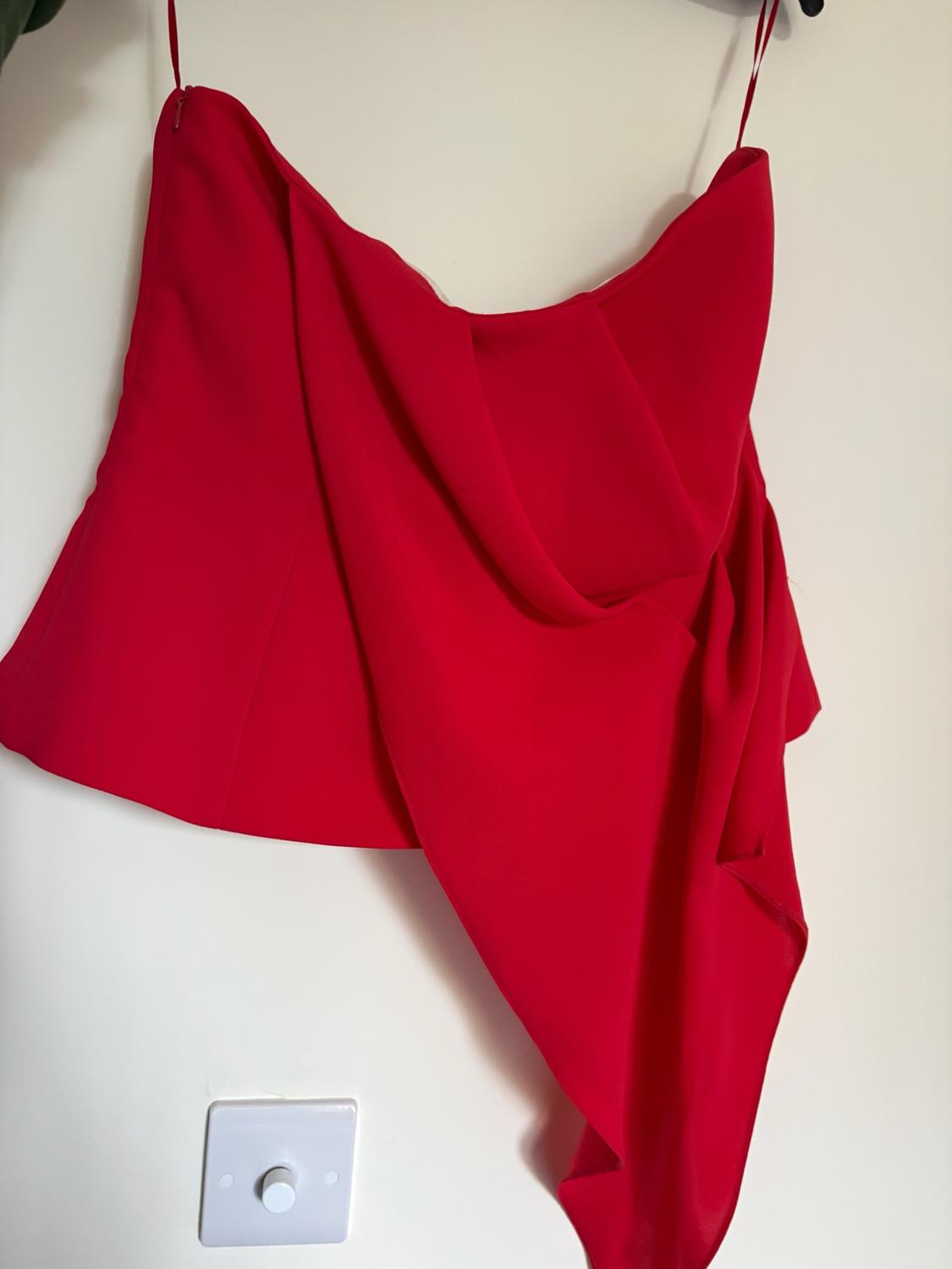 Asymmetric red camisole top with draped detail, hanging on a white wall above a light switch.