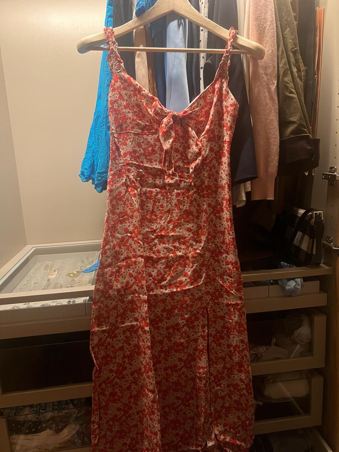 Red dress with a white floral print, shoulder straps and a bow on the chest, hanging on a wooden hanger in a wardrobe.