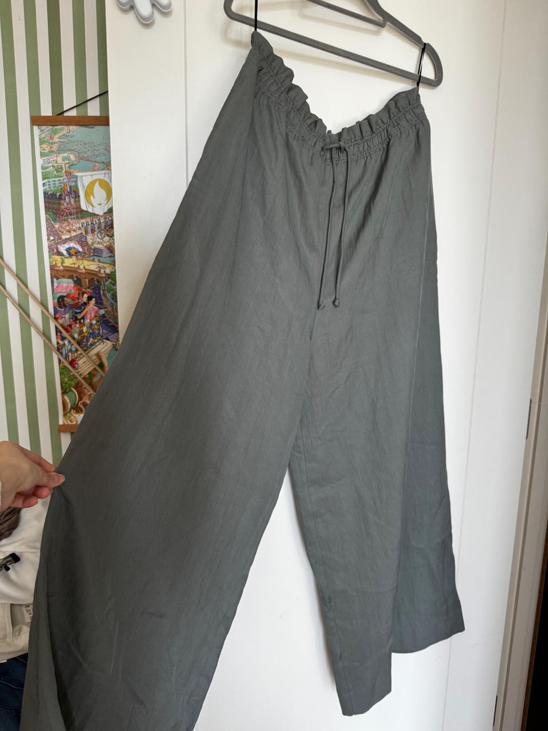 Grey wide-leg trousers hanging on a hanger, with an elasticated waist and drawstring, a background with green stripes and a colourful poster.