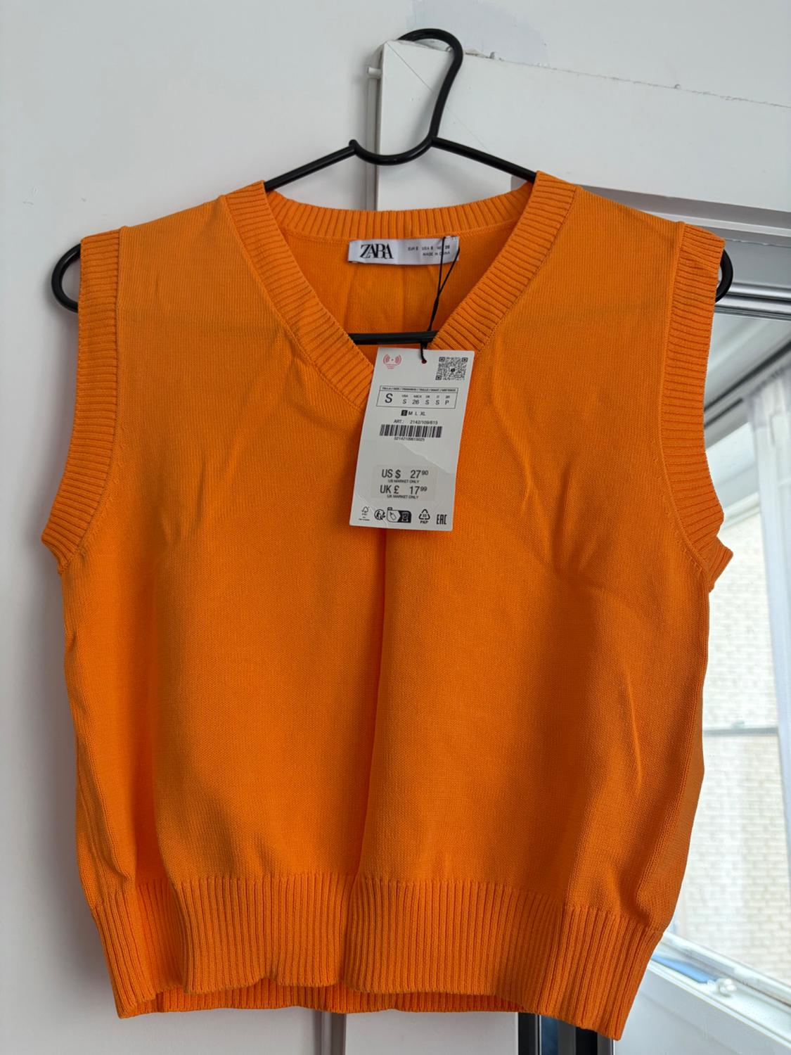 Orange knitted V-neck waistcoat hanging on a black hanger. Label with text: ZARA S US $ 27.90 UK £ 17.99.