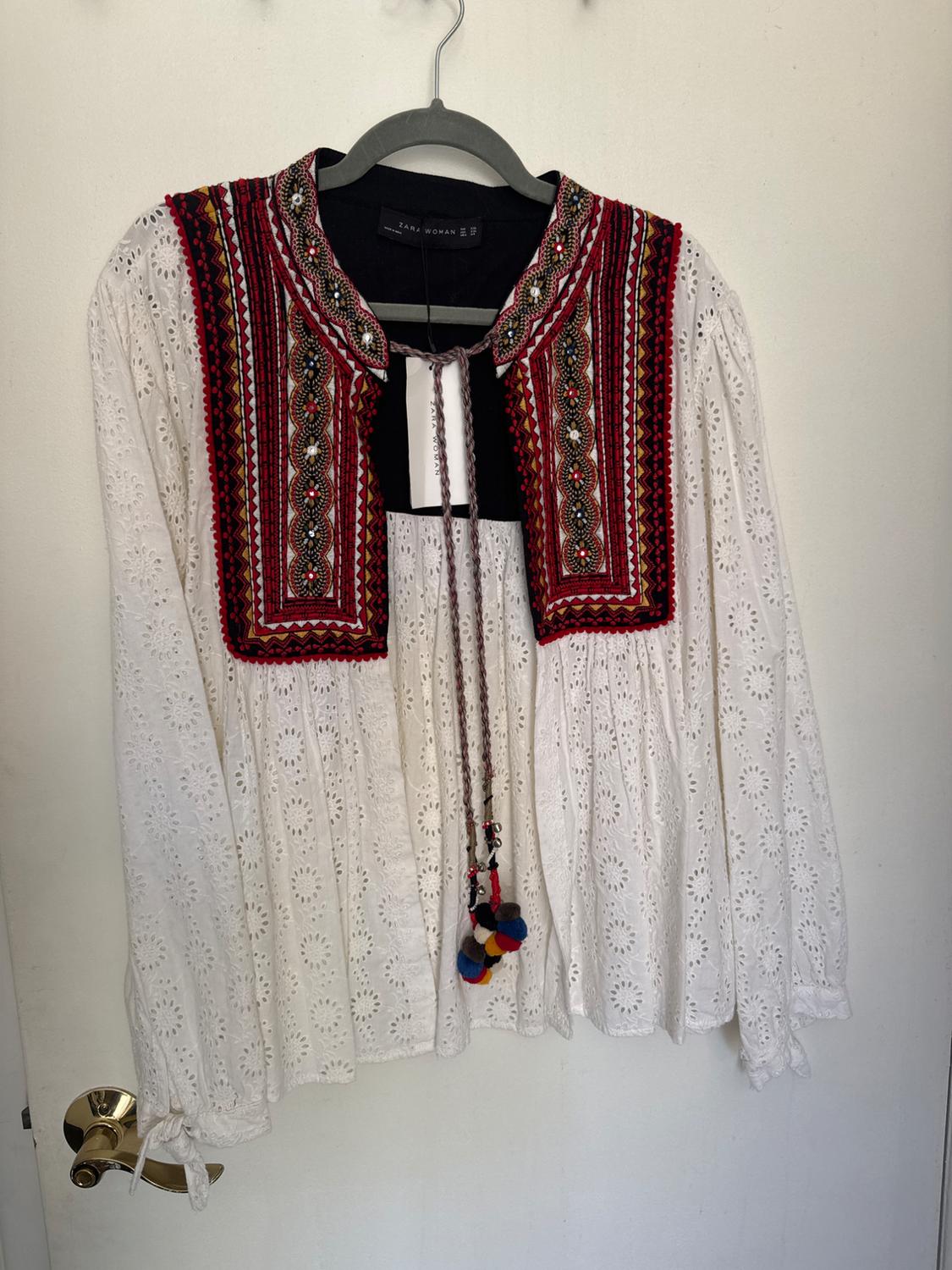 White lace blouse with red and black embroidery on the chest and collar, hanging on a hanger over a white door.