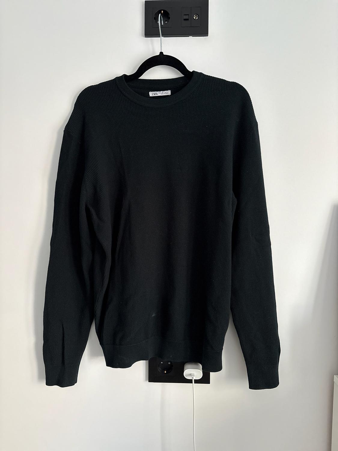 Black knitted jumper with long sleeves and a round neck, hanging on a black hanger against a white wall. The label reads ZARA.