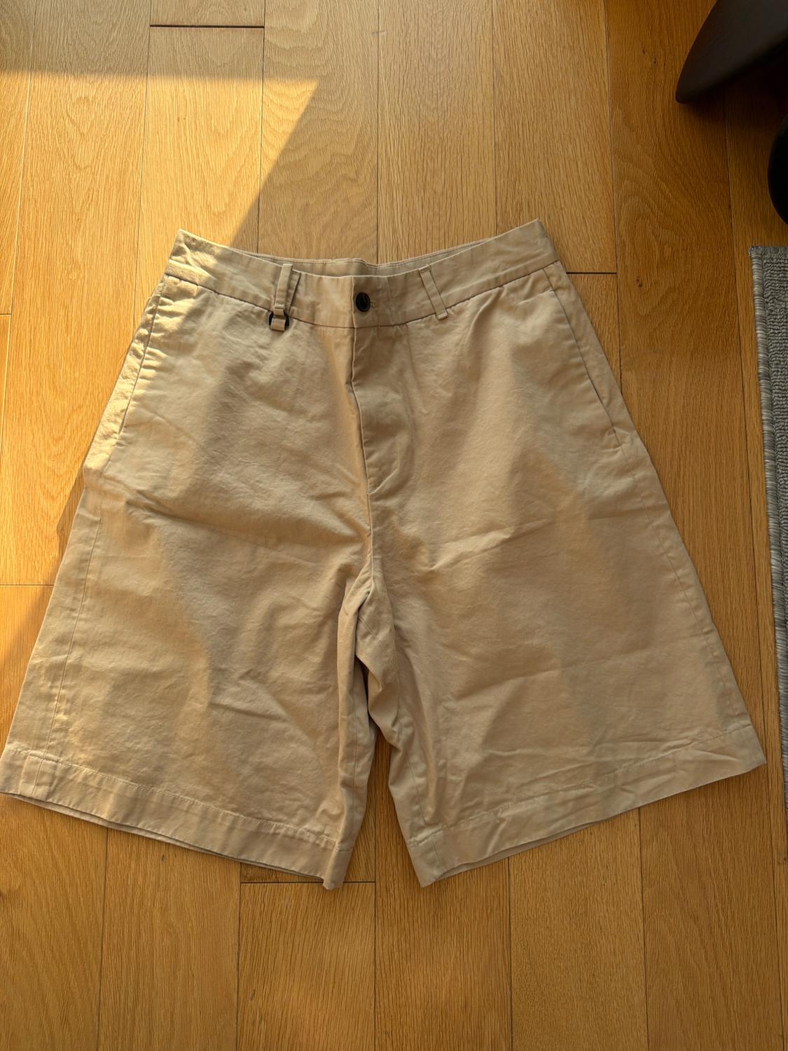 Light-fabric beige shorts with a black button, placed on a light wood floor, illuminated by natural light.