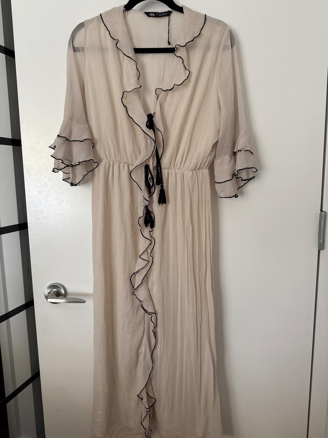Long beige dress with ruffles, black trim and tassels, on a black hanger against a white door. Relaxed style. Text: ZARA.