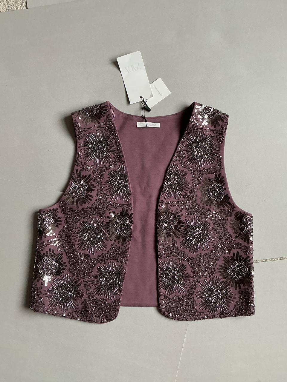 Lilac sleeveless waistcoat with floral sequin pattern on a grey background. Tags hanging from the neck.