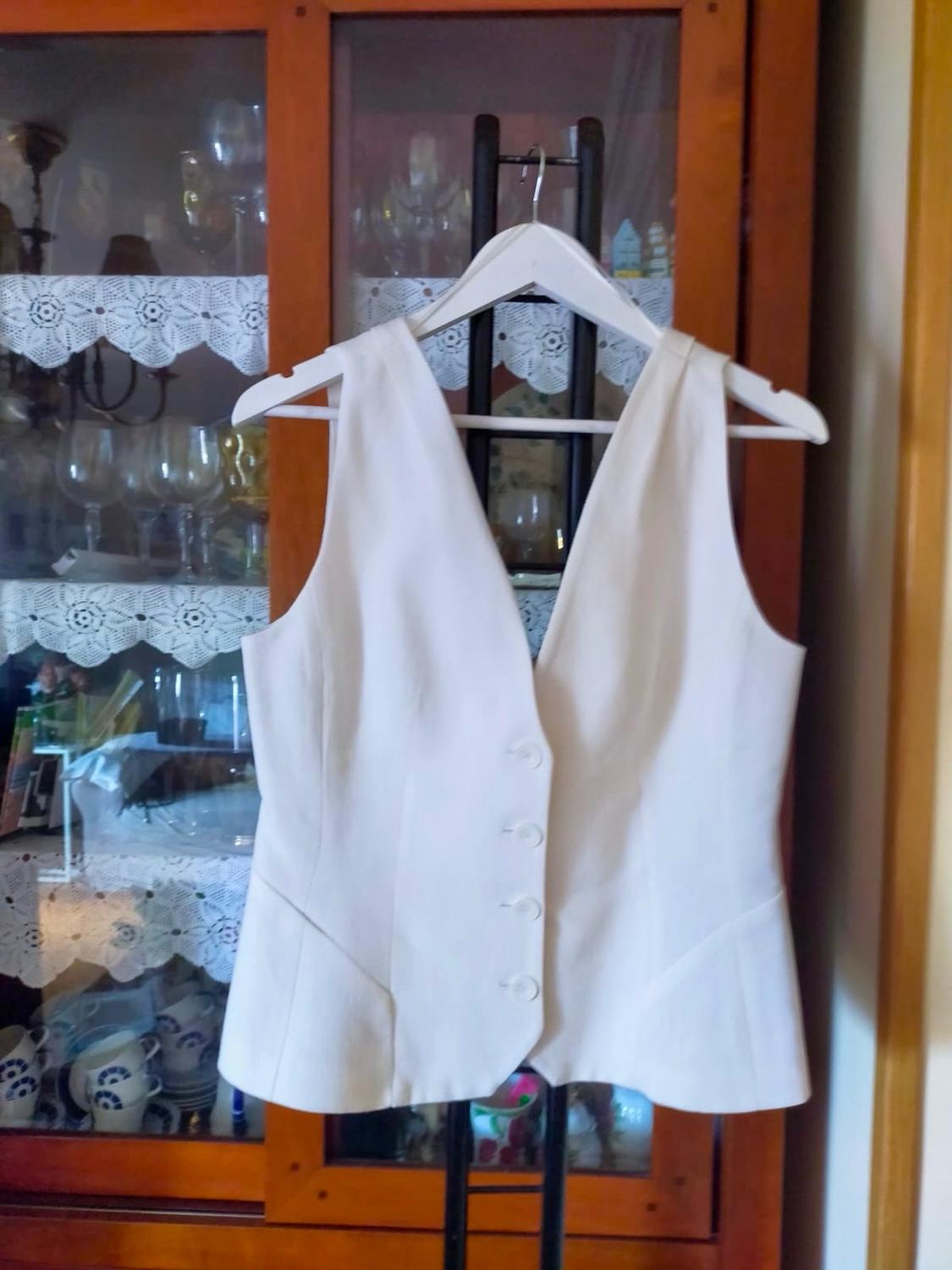 Buttoned white waistcoat with a V-neck, hanging on a white hanger in front of a wooden display cabinet with glassware.