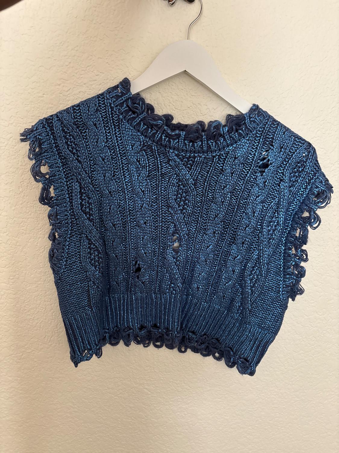 Blue knitted vest with a braided design and decorative edges, hanging on a white hanger against a beige wall.