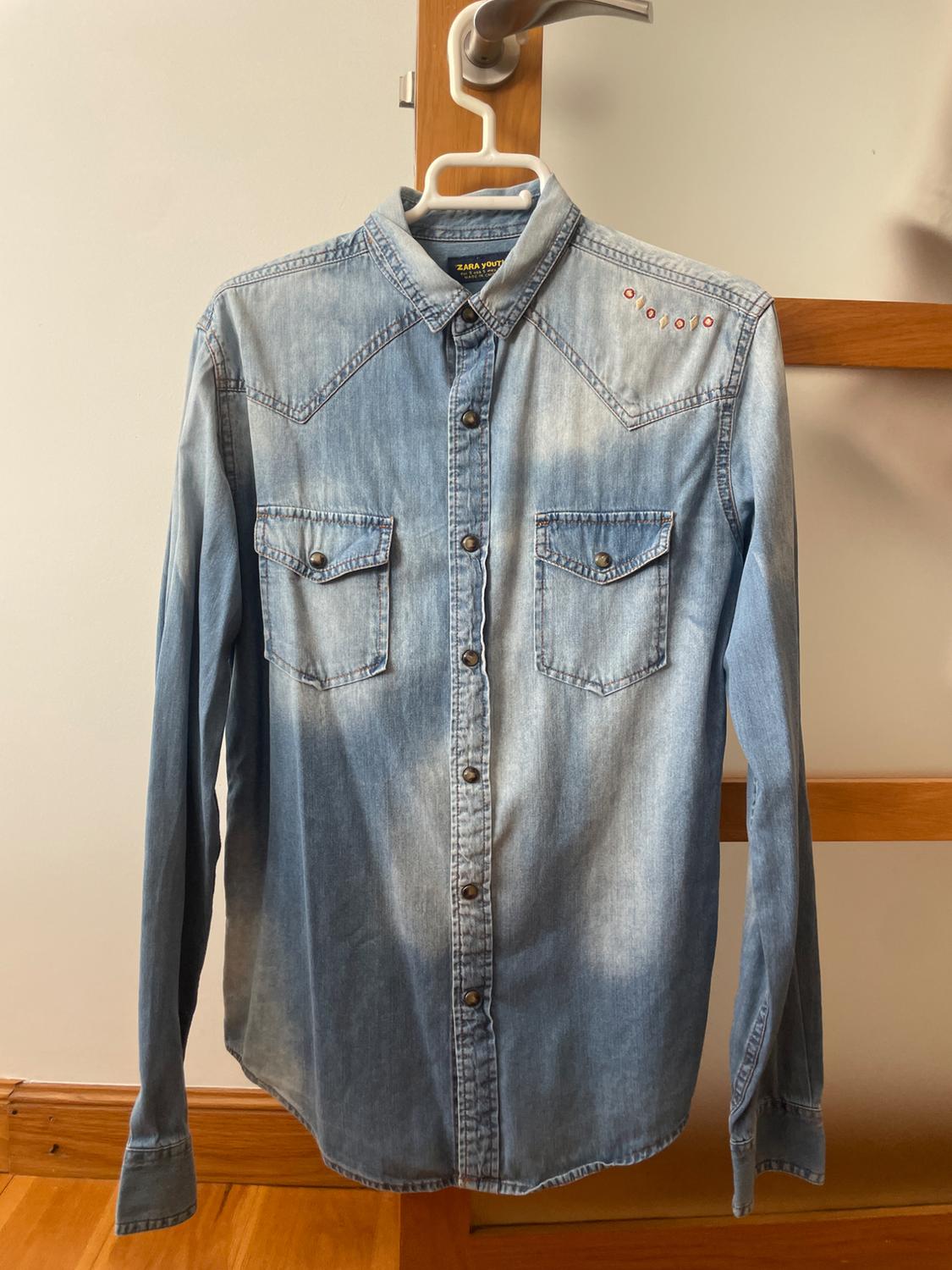 Light blue denim shirt hanging on a hanger. It features a washed effect, chest pockets and a neck label with the text ZARA YOUTH.