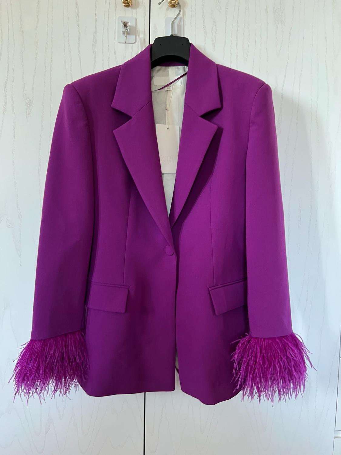 Purple one-button blazer with flap pockets and feathers on the cuffs, hanging on a hanger in front of a white door.