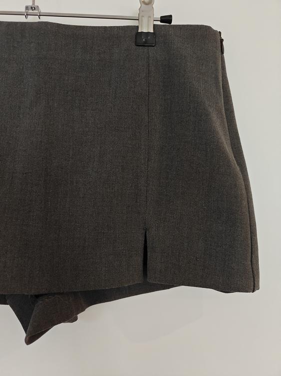 Grey short skirt hanging on a white hanger against a white background. The fabric has a smooth texture and a small front pleat.