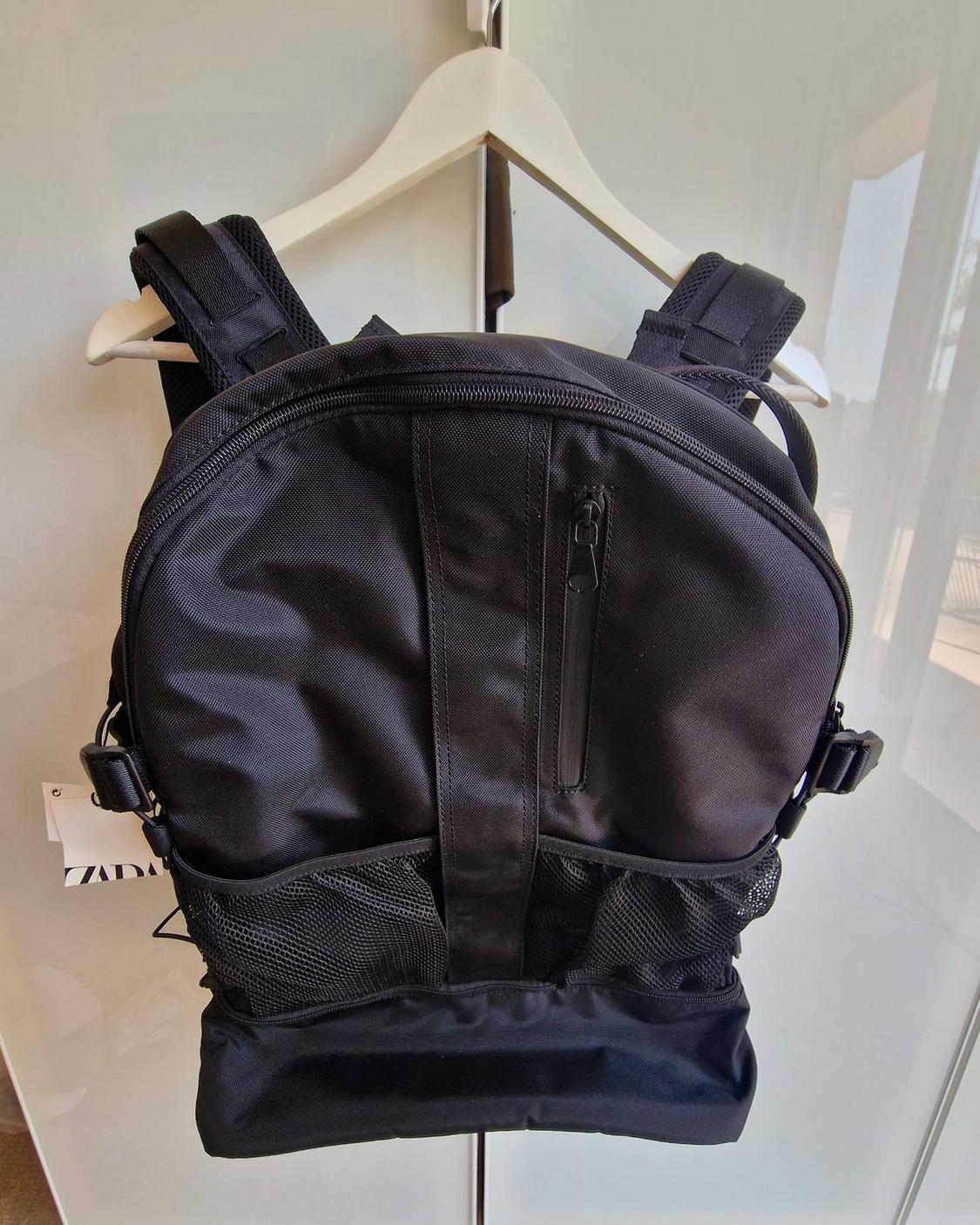 Black backpack hanging on a white peg. It features mesh pockets, a front zip and a side label with the text ZARA.
