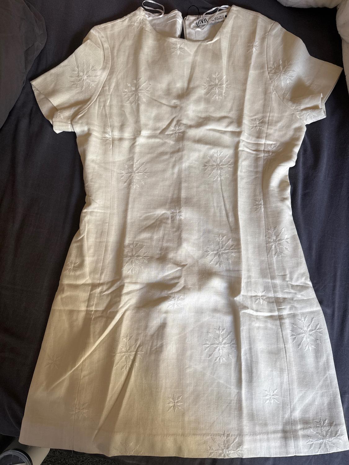 Beige short-sleeved dress with floral embroidery, on a dark background. Visible zip on the upper back.