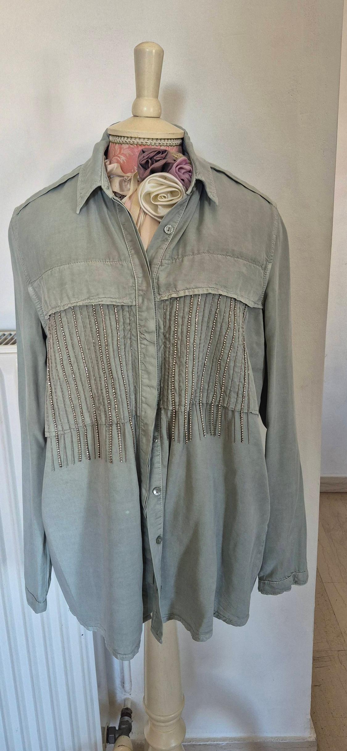 Mannequin in a light green shirt decorated with beaded fringes on the chest. White background and floral details on the collar.