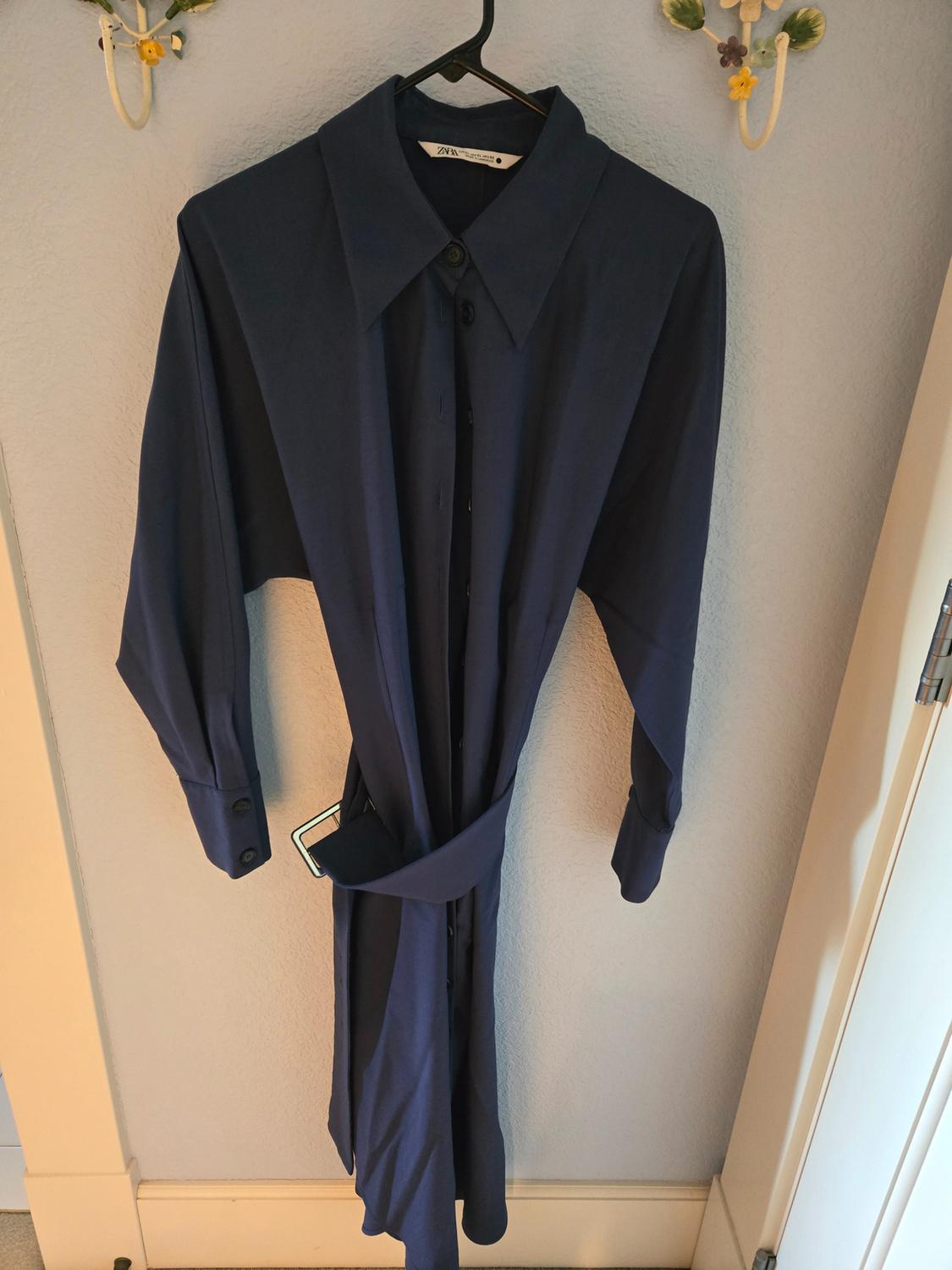 Dark blue dress hanging on a hanger, with a wide collar and matching belt. Light wall background with floral decoration.