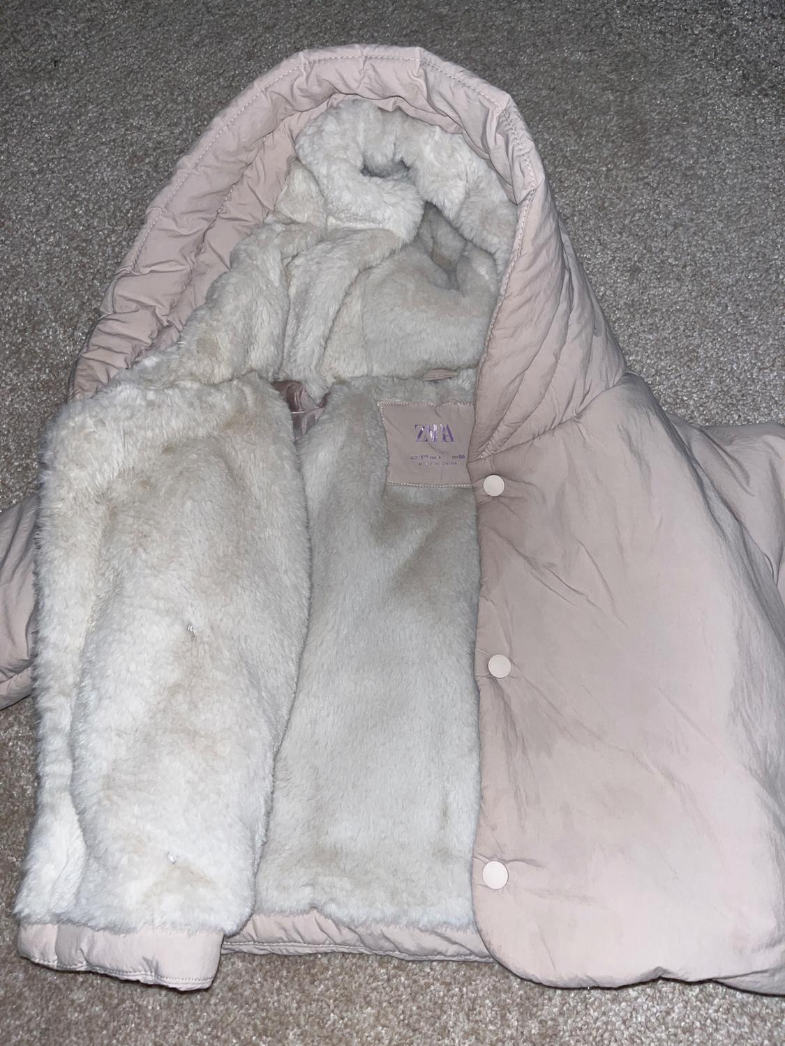 Light pink coat with white plush interior, hood, and snap buttons, on a beige carpet. Visible label with text "ZARA".