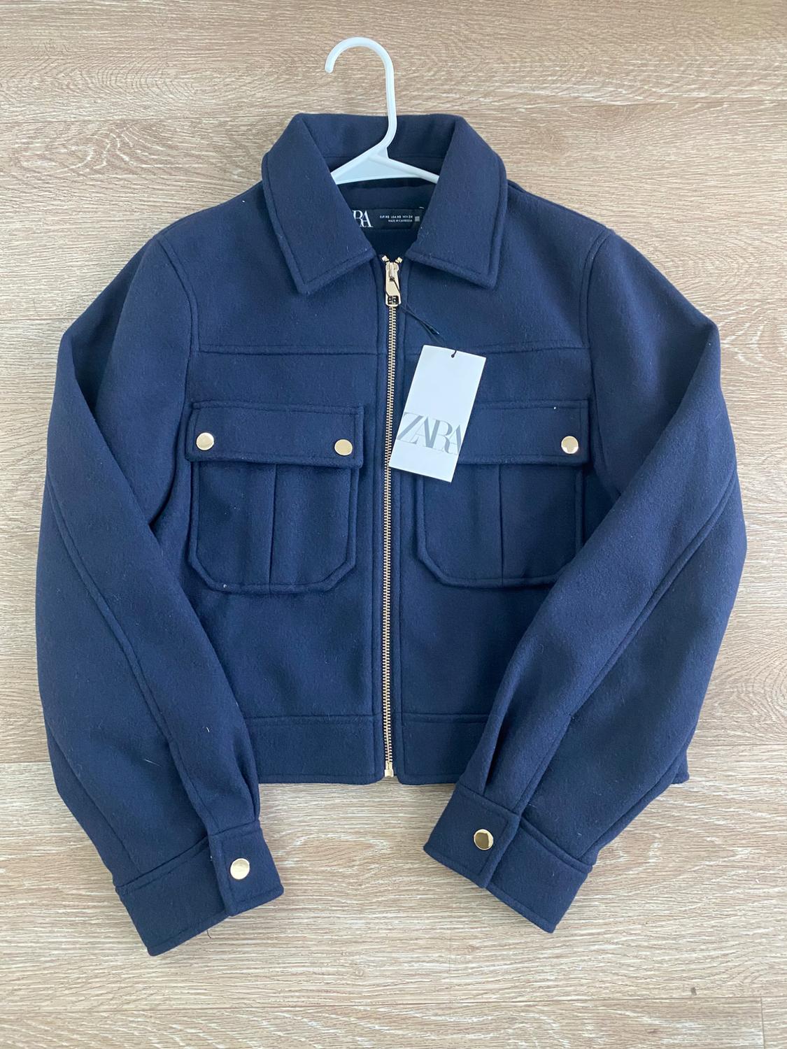 Navy blue jacket with a gold zipper and front pockets, hanging on a white hanger against a light wood background. "ZARA" tag.