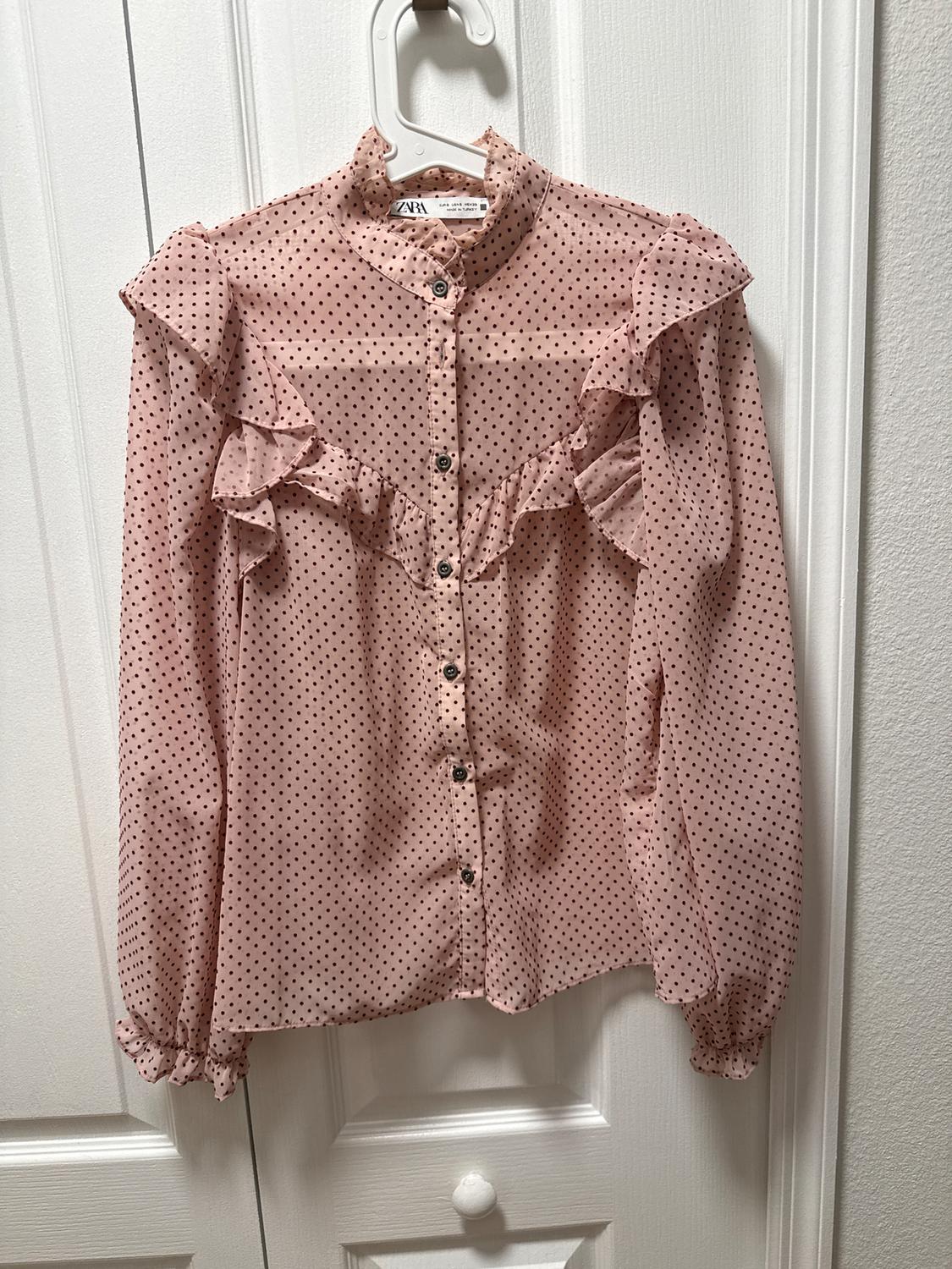 Pink blouse with black polka dots and ruffles on the shoulders, hanging on a white hanger in front of a white door.