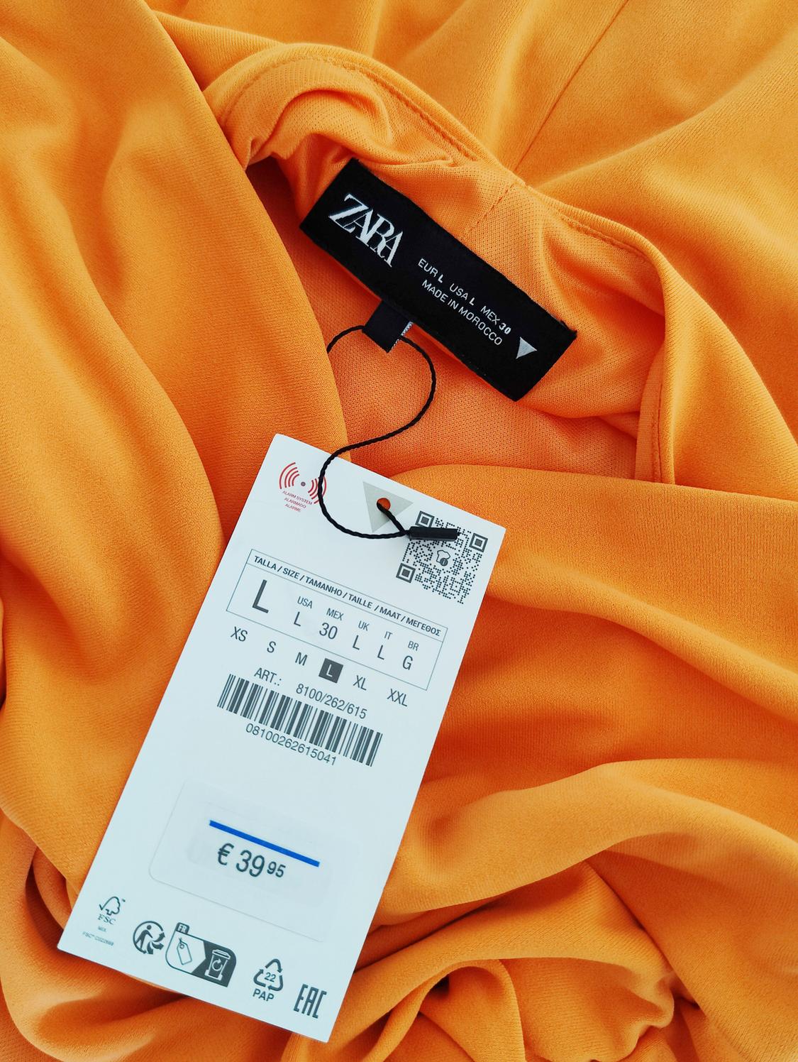 Ετικέτα Zara σε πορτοκαλί ύφασμα, εμφανίζει μέγεθος L, τιμή €39,95, γραμμωτός κώδικας, QR και λεπτομέρειες κατασκευής στο Μαρόκο.
