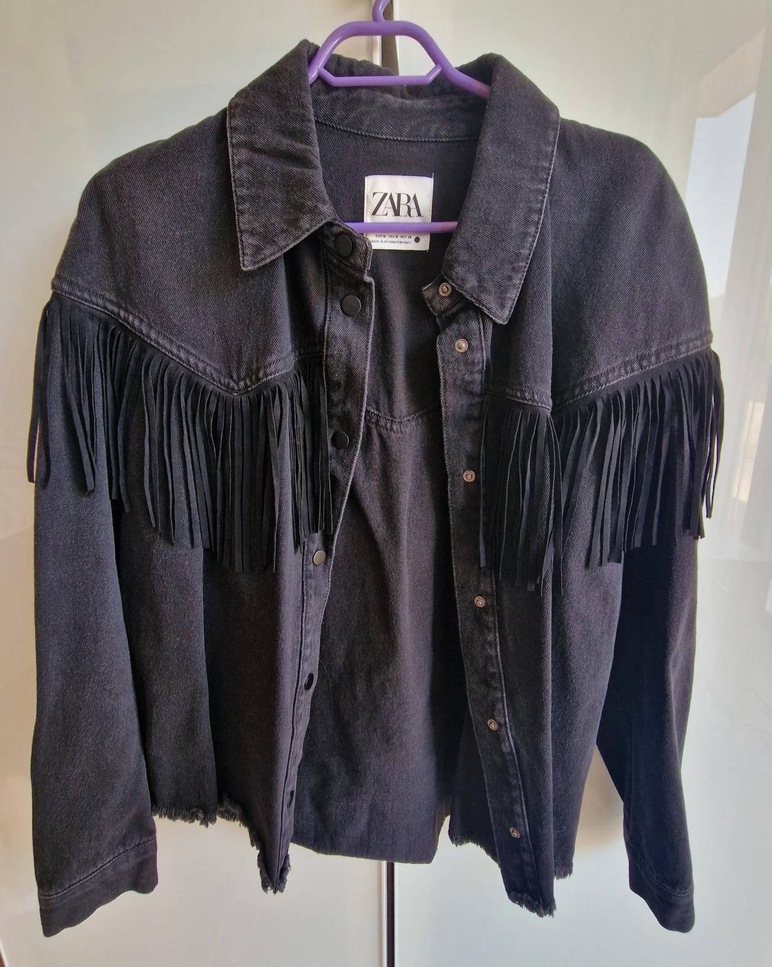 Black denim jacket with fringing on the chest and sleeves, hanging on a purple hanger. Inner label bearing the text ZARA.