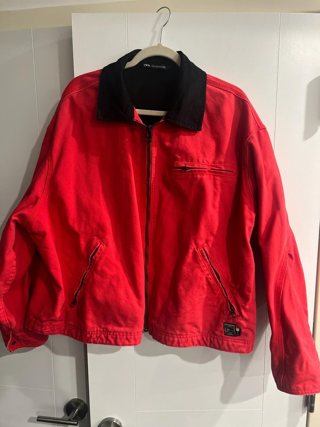 Red jacket with a black collar and zips, hanging on a hanger on a white door. Label with the text ZARA.