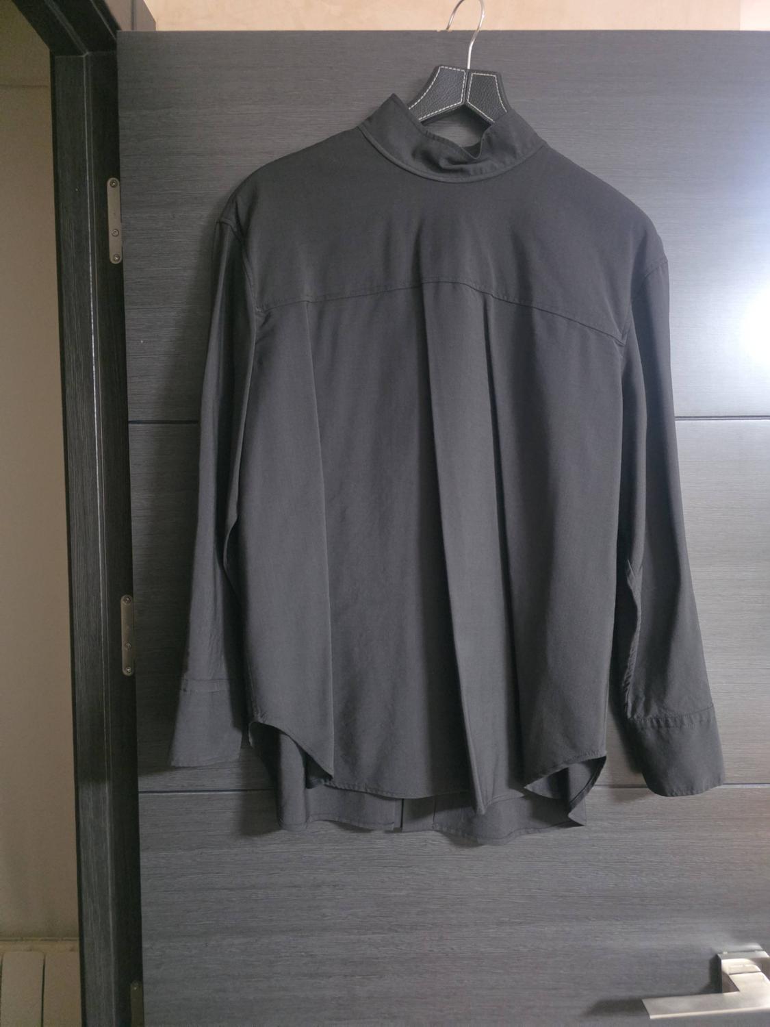 Dark grey long-sleeved shirt hanging on a hanger on a wooden door. Back view with a high collar and pleat.