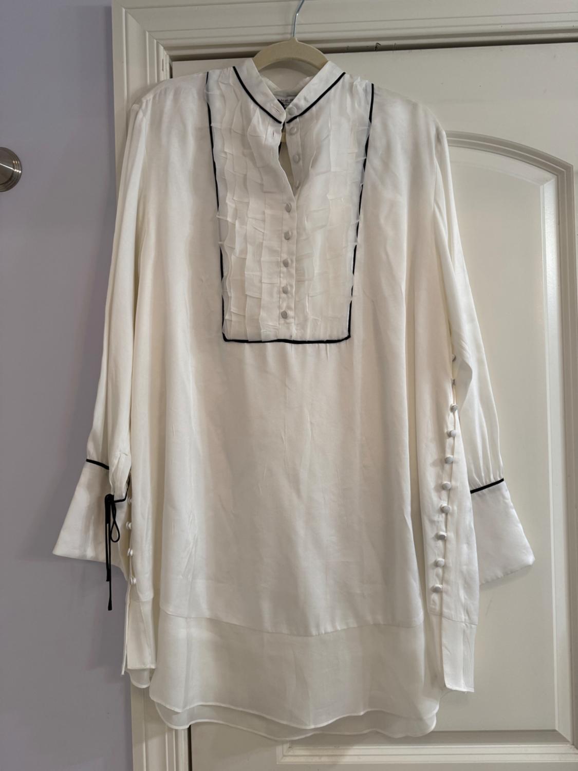 White blouse hung on a hanger, with pleated details on the front and black trim on the collar and sleeves. White door background.