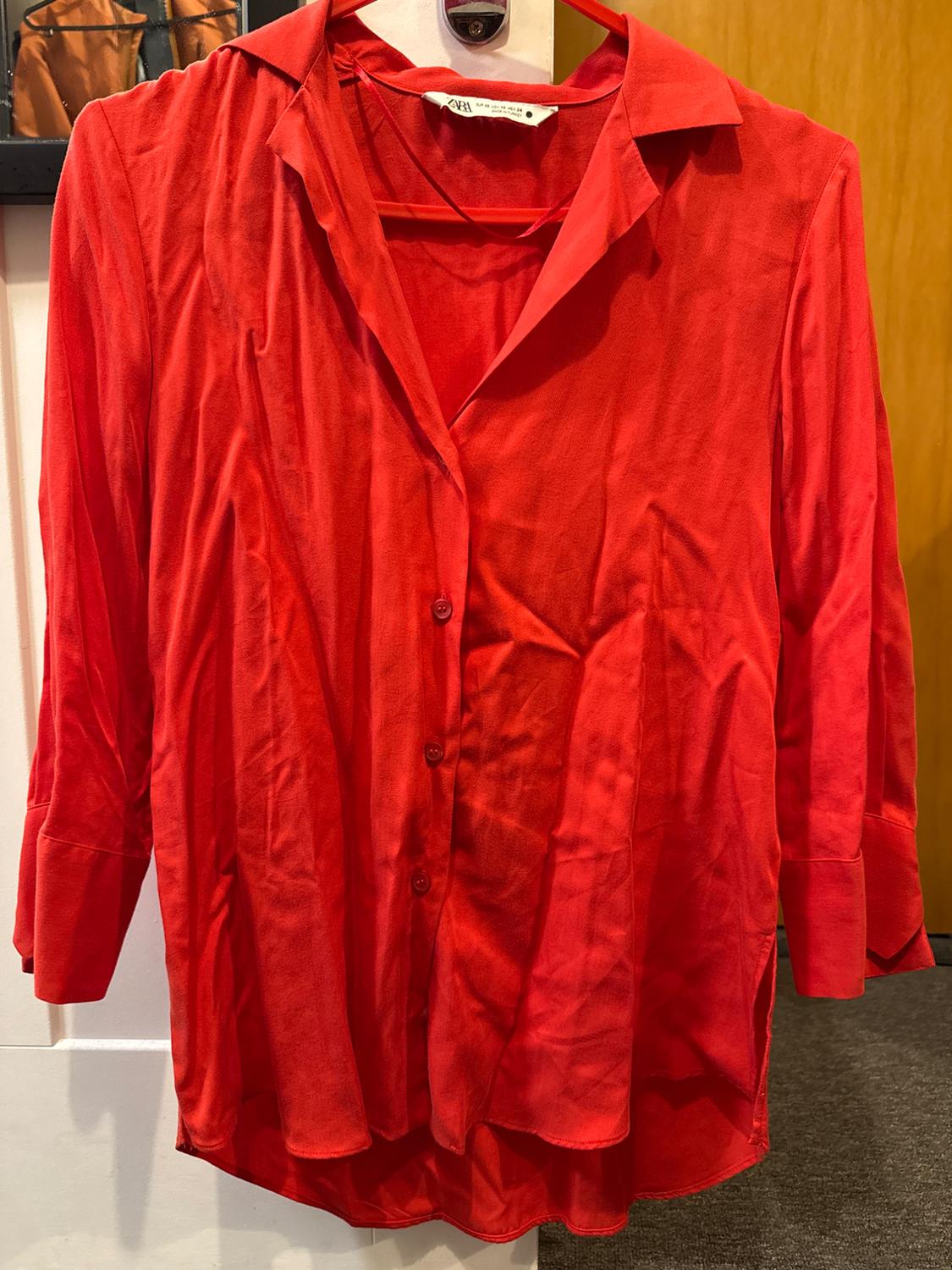Long-sleeved red shirt hanging on a hanger, with a classic collar and front buttons. White wall background and wooden door.