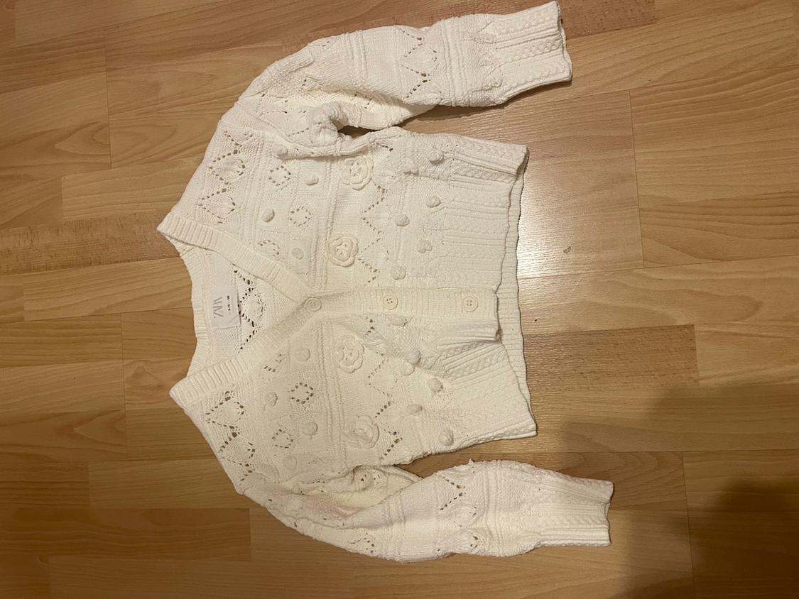 White knitted cardigan with bobble details and openwork patterns, on a light wooden floor. Visible label: ZARA.