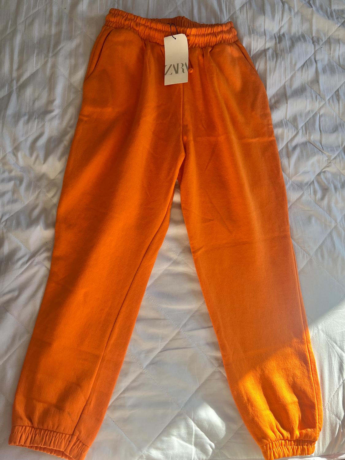 Orange trousers with an elasticated waist and a "ZARA" label, on a white quilt with a diamond pattern.