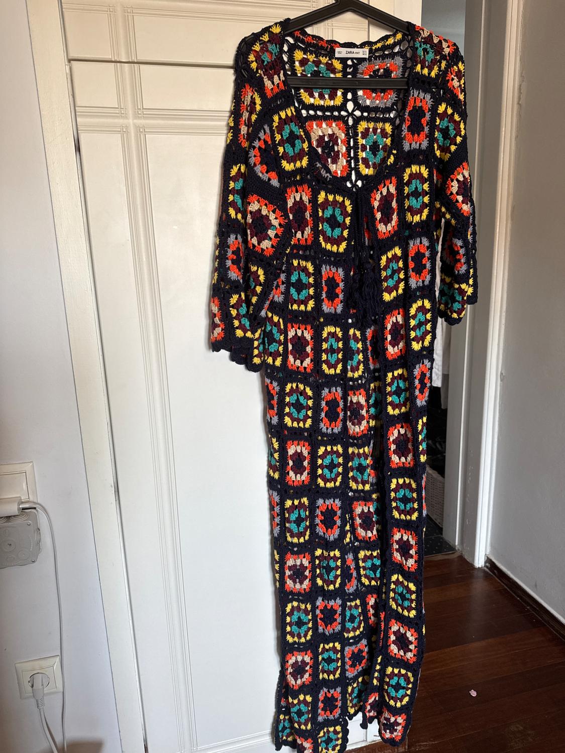 Crochet dress hanging on a hanger, with a colourful chequered pattern in red, blue, yellow, and green on a black background.