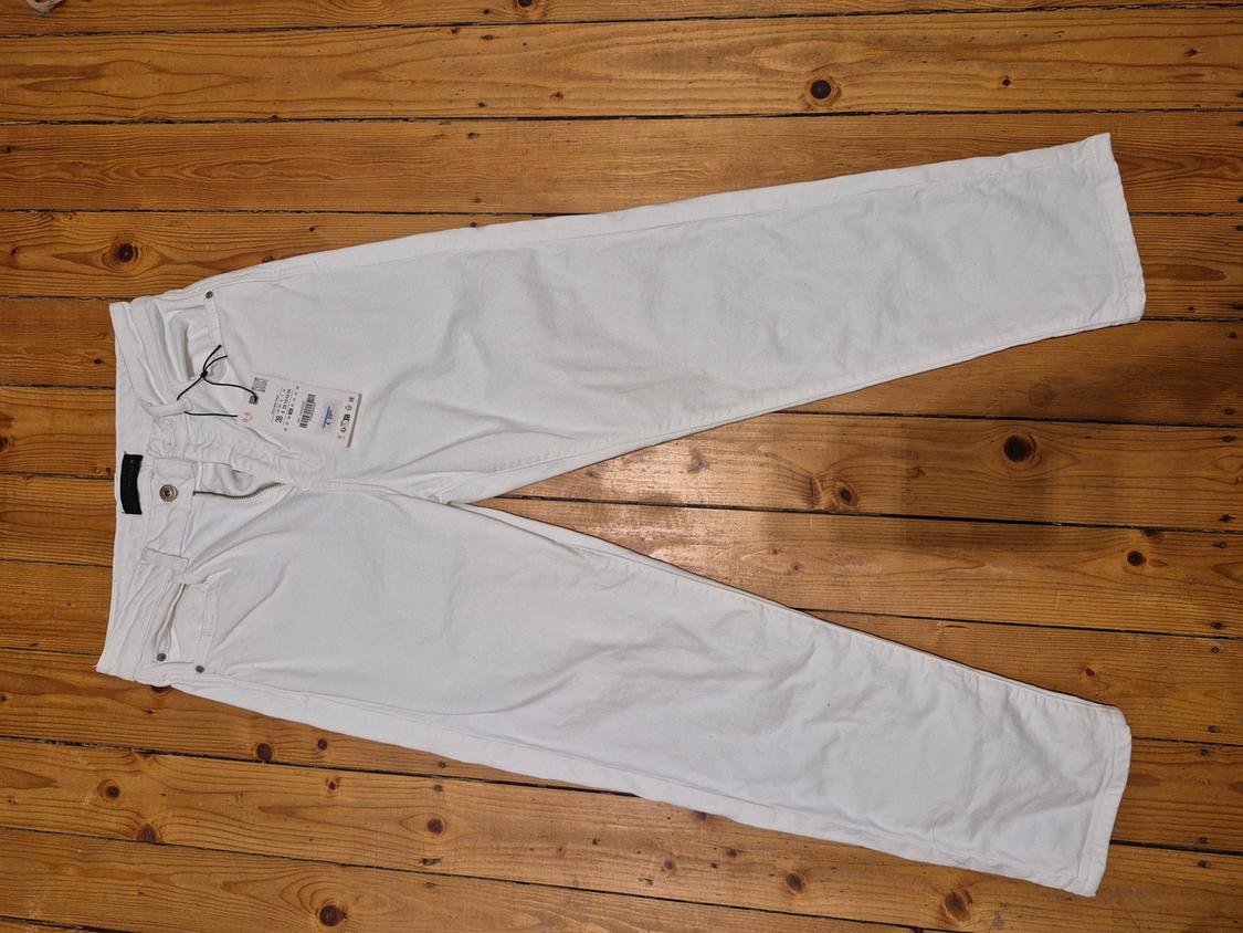 White trousers laid out on a wooden floor, with a visible swing tag on the waistband.