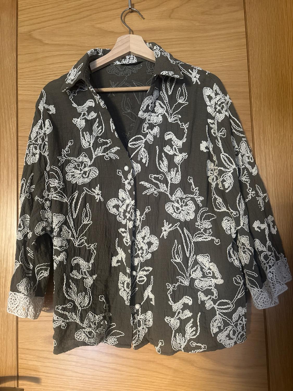 Dark green button-up shirt with white floral embroidery and lace cuffs, hanging on a hanger against a wooden background.