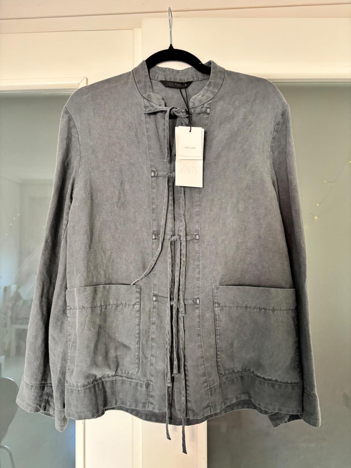 Grey linen jacket hanging on a hanger, with two large front pockets and a visible label that reads "100% Linen".