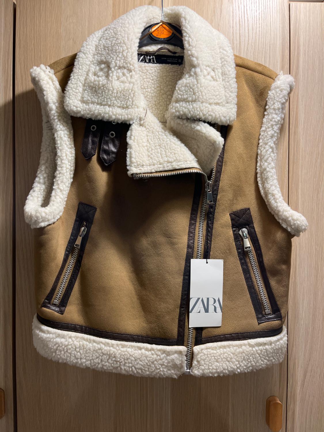 Brown gilet with a white fur lining and zip fastenings, hanging in a wooden wardrobe. Label with the text ZARA.