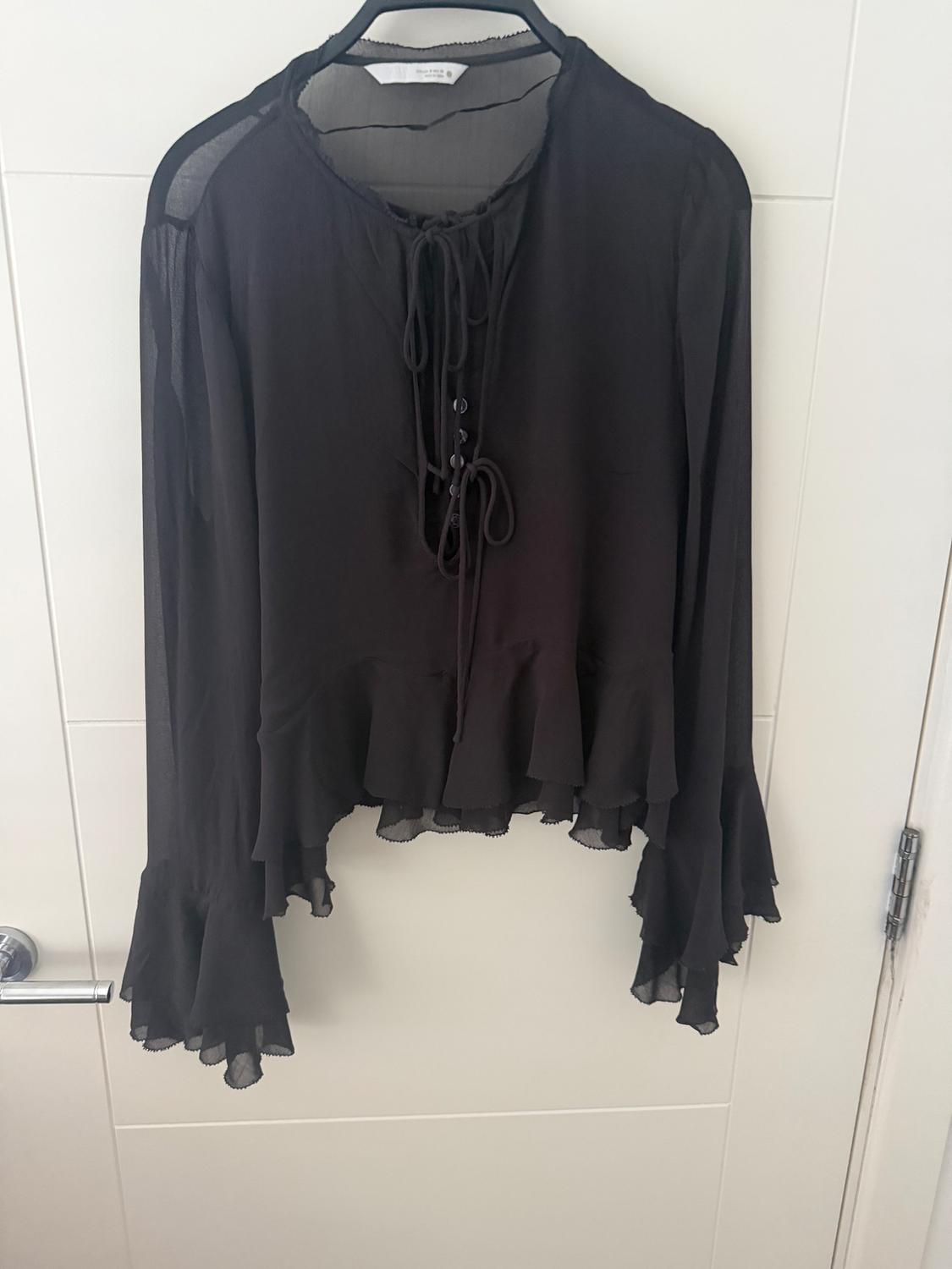 Black chiffon blouse with long flared sleeves and lacing at the front, hanging on a hanger over a white door.