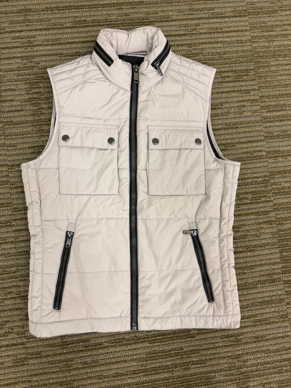 Light beige quilted vest with a high neck, black center zipper, two chest pockets with a flap, and two zippered pockets.