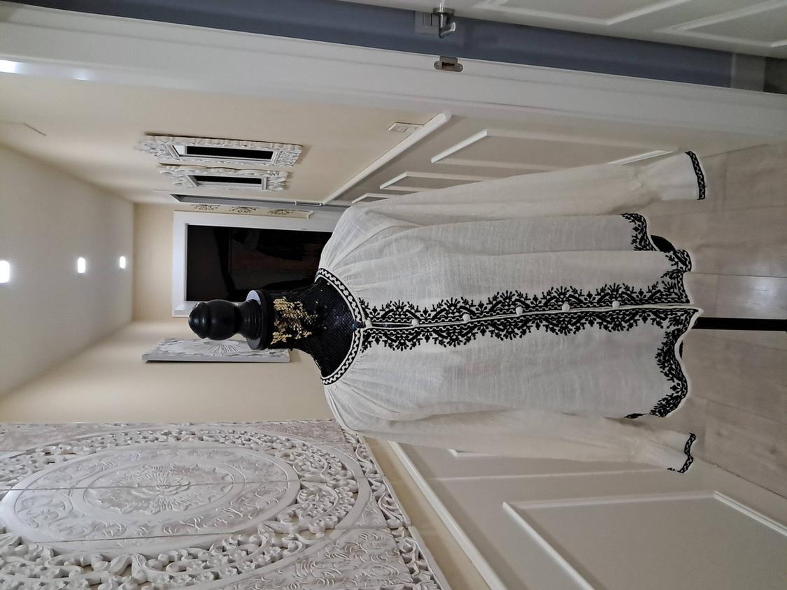 Mannequin in a white long-sleeved blouse with black embroidery in an illuminated hallway, decorated with carved wall panels.