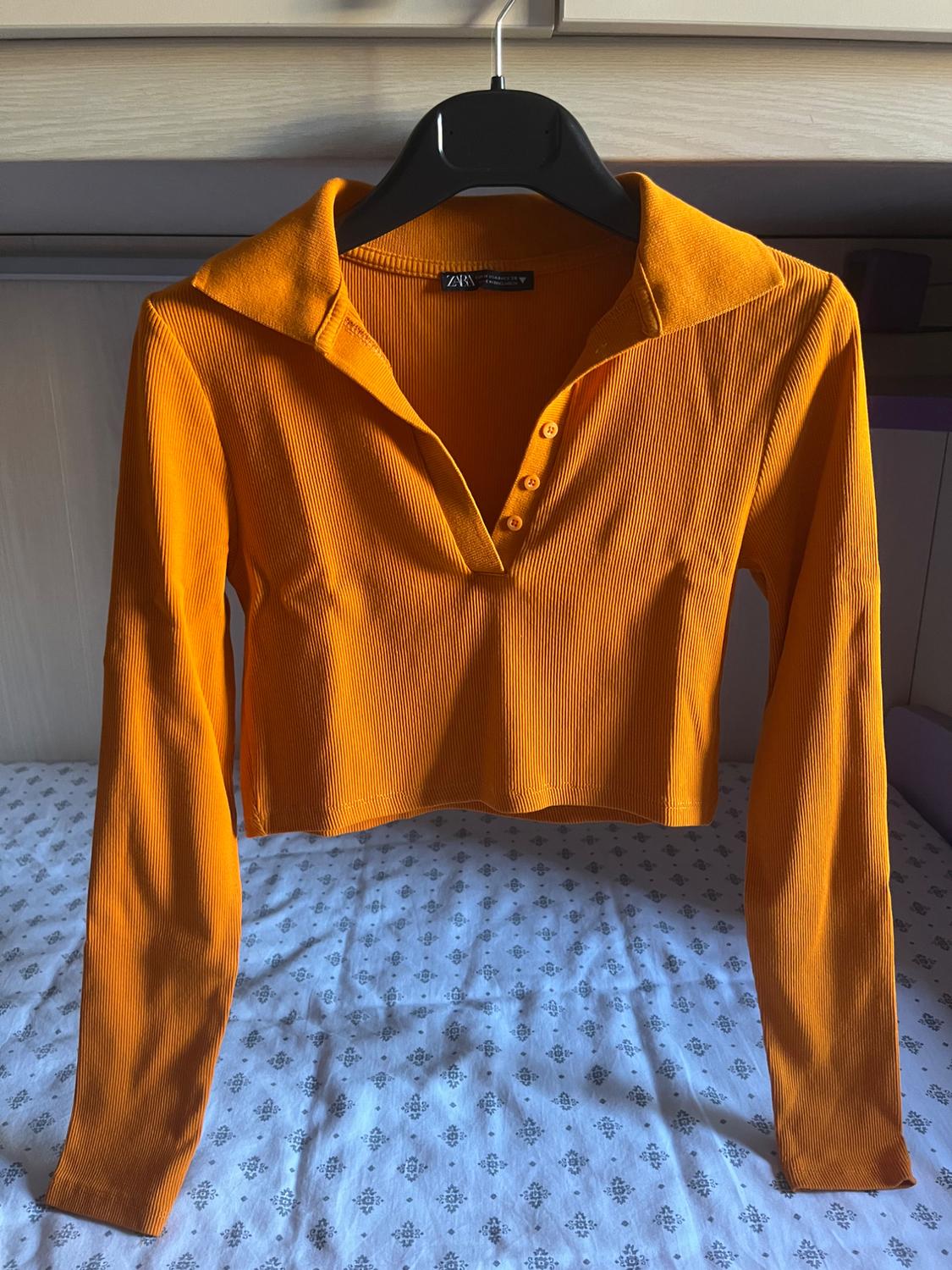 Orange ribbed long-sleeved, collared, and button-up crop top, with ZARA tag. Hung on a hanger over a bed with patterned sheets.