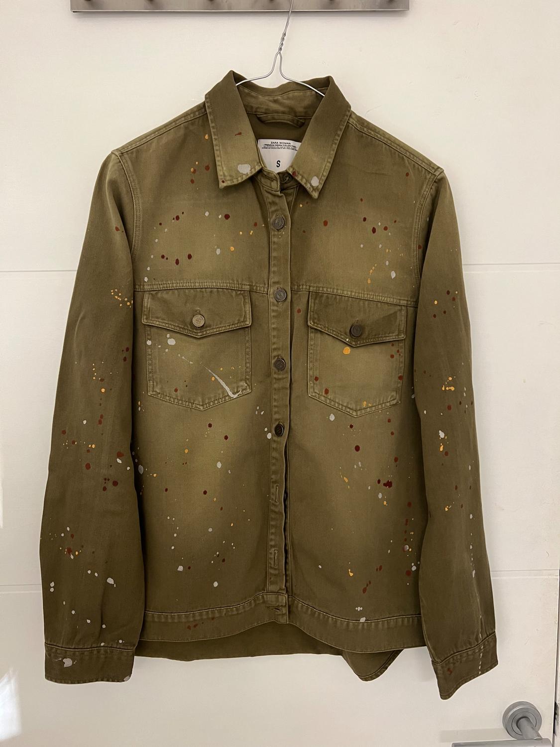 Olive green jacket with multicoloured paint splatters, hung on a metal hanger against a white background.
