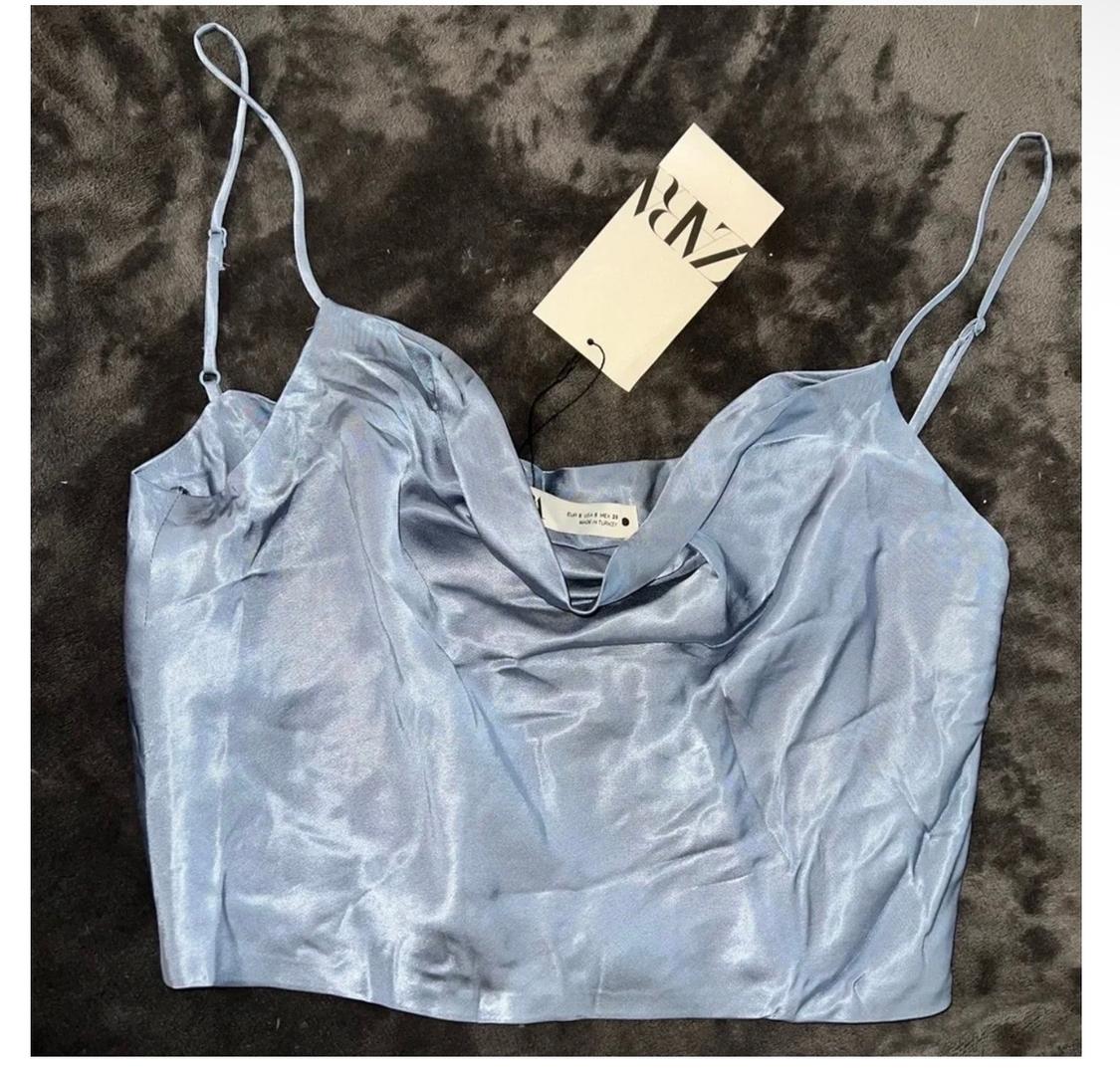 Light blue spaghetti strap top on a dark grey background, with a hanging "ZARA" tag. Fabric with a subtle sheen.