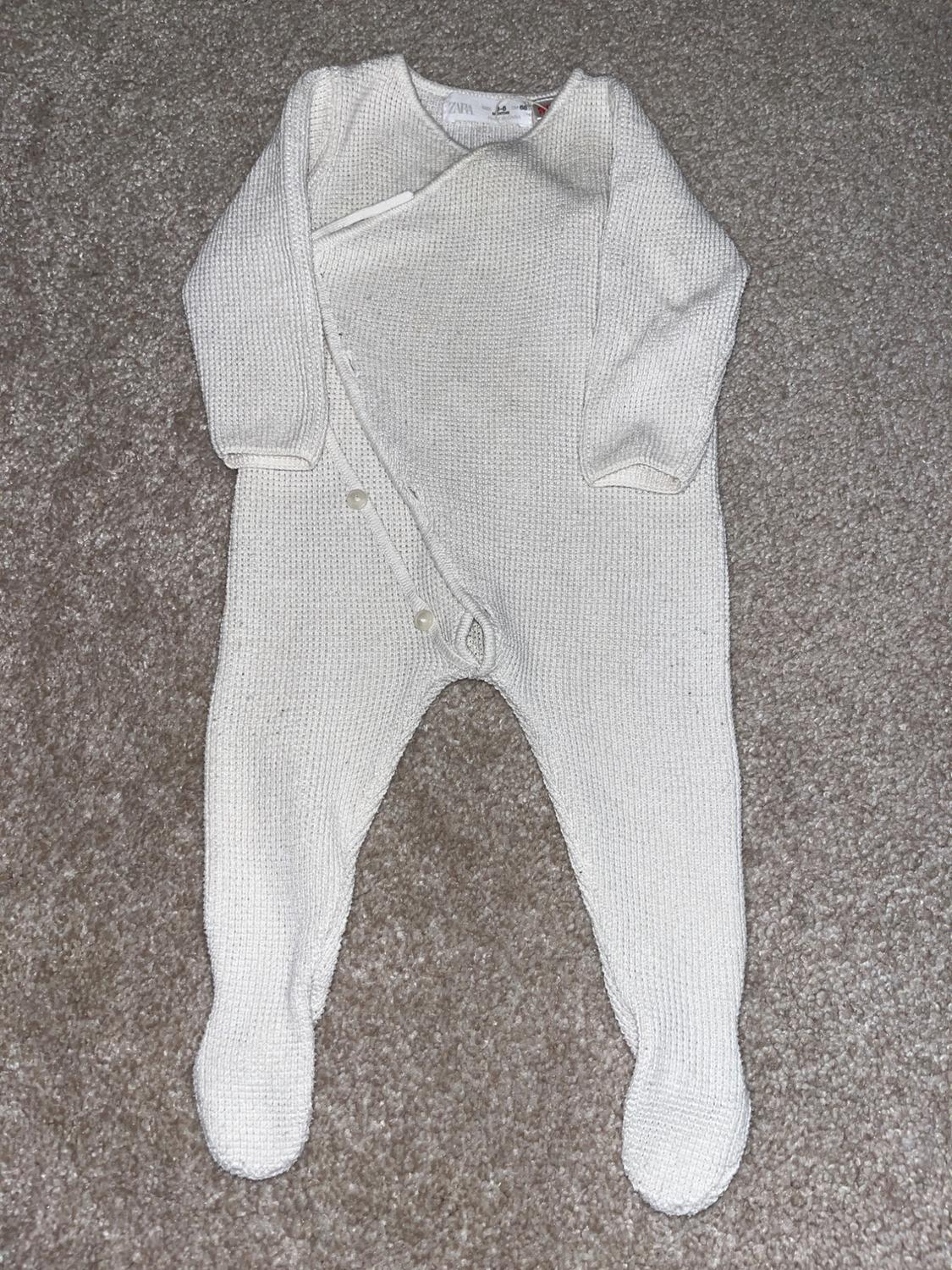 White knitted romper with buttons on the front, on a grey rug. Visible label on the collar.