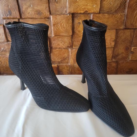 Black mesh high-heeled ankle boots on a white surface, with a geometric textured wooden wall in the background.