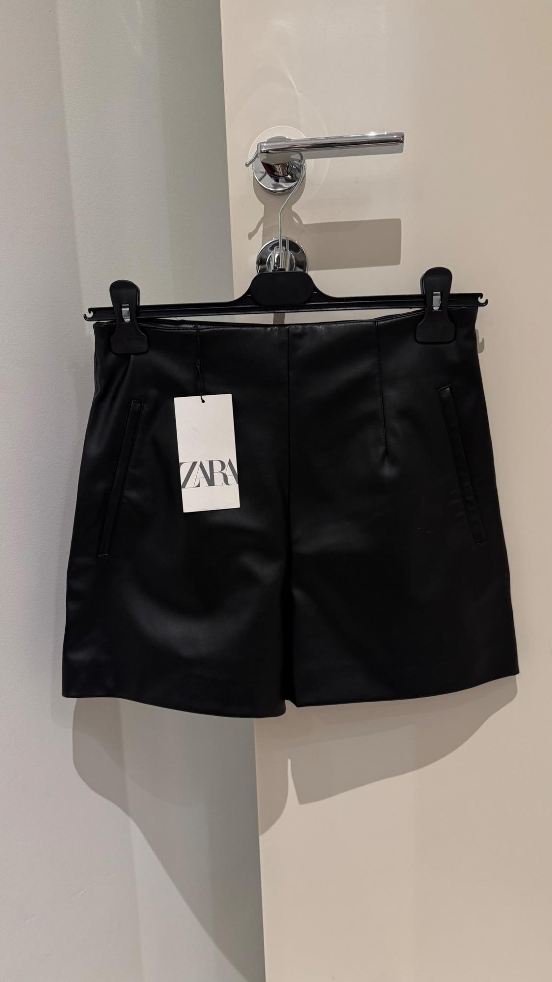 Black shorts hanging on a hanger with a label that says "ZARA", over a white door with a silver doorknob.